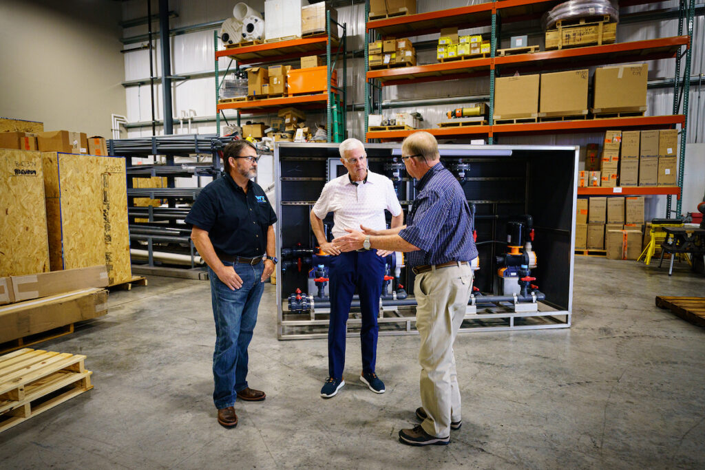 Three male Water Tech employees wearing safety glasses having a conversation in a warehouse.