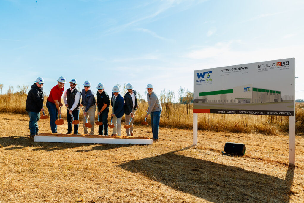 The Water Tech team with white hard hats shoveling brown dirt and smiling at the camera beside the sign depicting their new North Carolina Distribution Center.