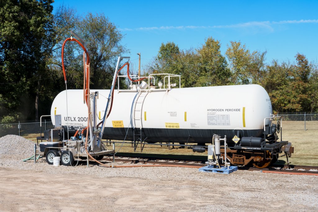 A long white hydrogen peroxide tank on a portable transloader. It is a sunny day and there is a long red hose running from the top of the tank.