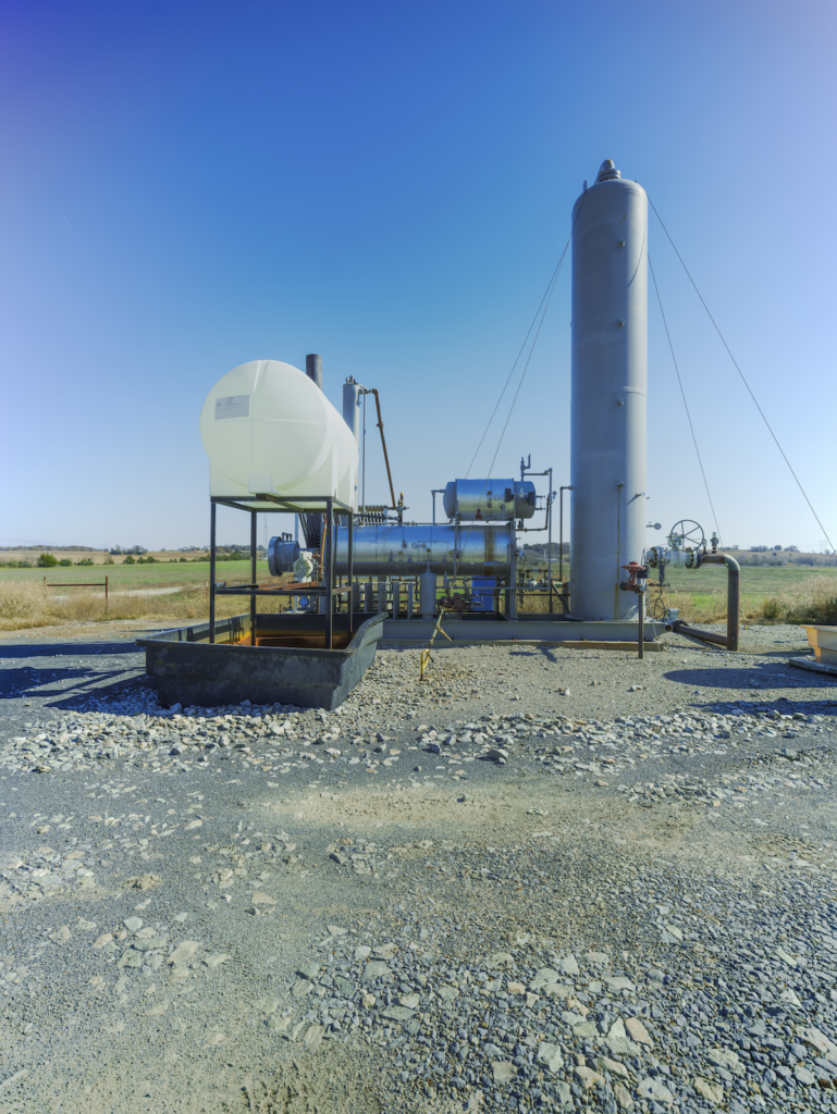 A gas dehydration unit and a white translucent salt water tank sitting in a gravel lot beside a field above a salt water well. The gas dehydration unit is tall and metal.