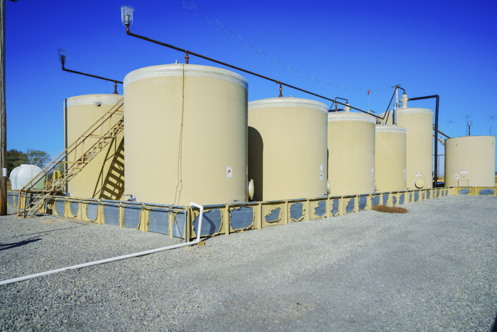 Two rows of large concrete-colored storage tanks in a pen. Piping runs along the top of the tanks and a rusty metal ladder runs up to the top of the center of the rows.