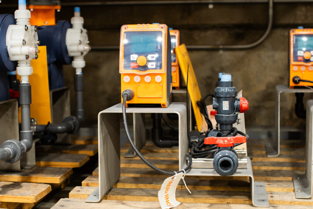 A bright orange chemical metering pump in the Water Tech warehouse. The gauge is small with multiple buttons and hooks up to the black pump.