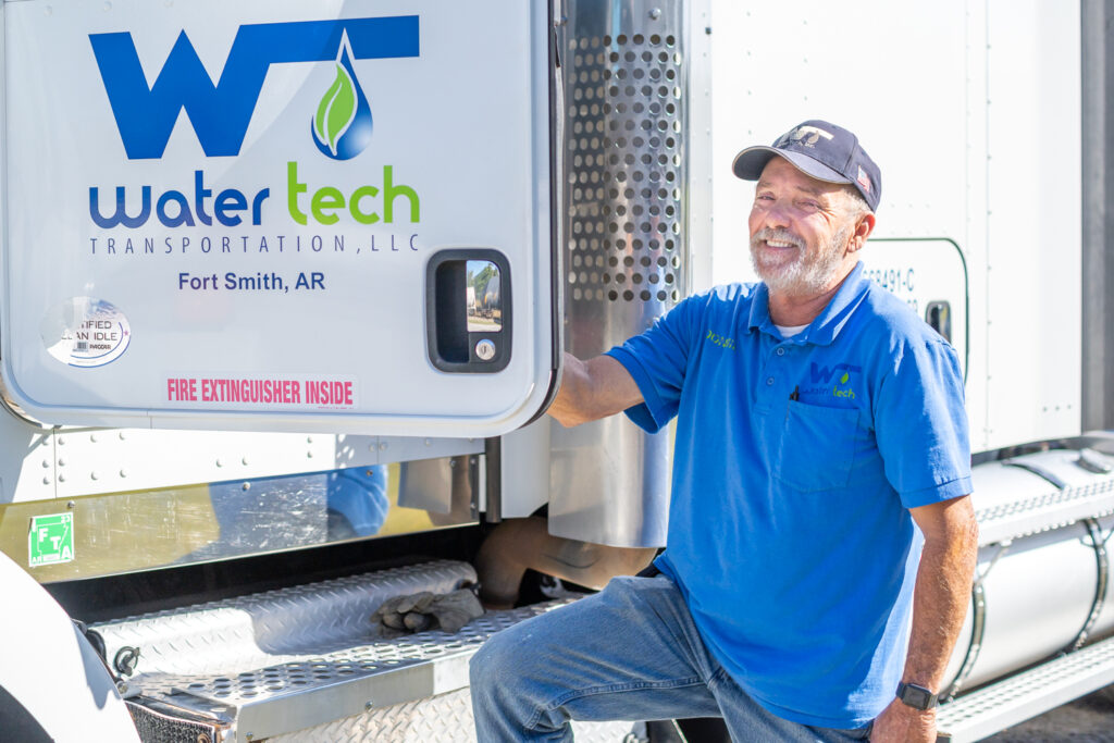 A Water Tech truck driver in a bright blue polo smiles for the camera as he holds open the door to the semi-truck. The door has the Water Tech logo on it.