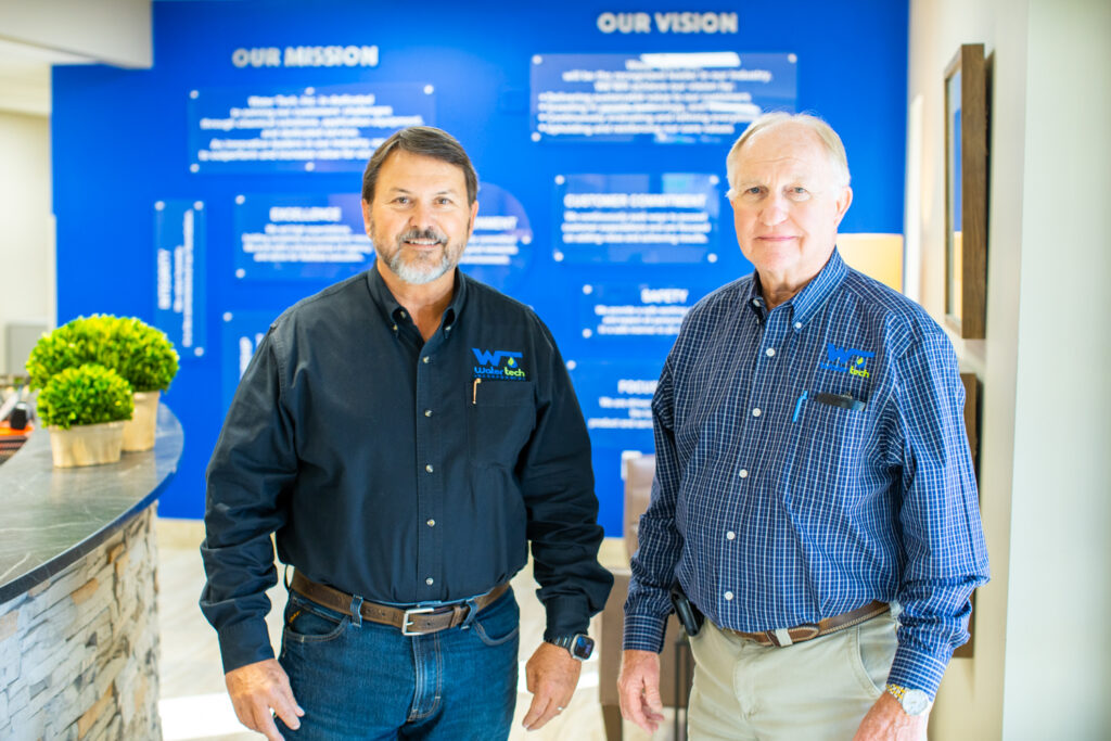 Two Water Tech employees standing in front of the bright blue Mission and Vision board. The men are older and one is wearing a black water tech button down and the other a blue plaid.