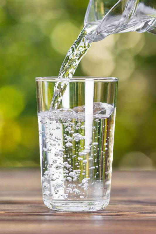 The tip of a glass pitcher pouring water into a clear glass cup. The cup is sitting on a wood table and the background is a blur of green.