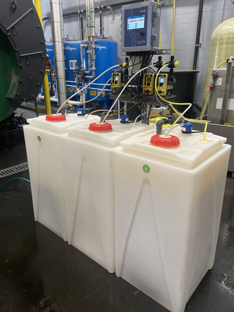 Industrial strength white-plastic chemical storage tubs. Three tubs sit together. There is a bright red cap on the top with yellow tubing running out of it and into a digital system.