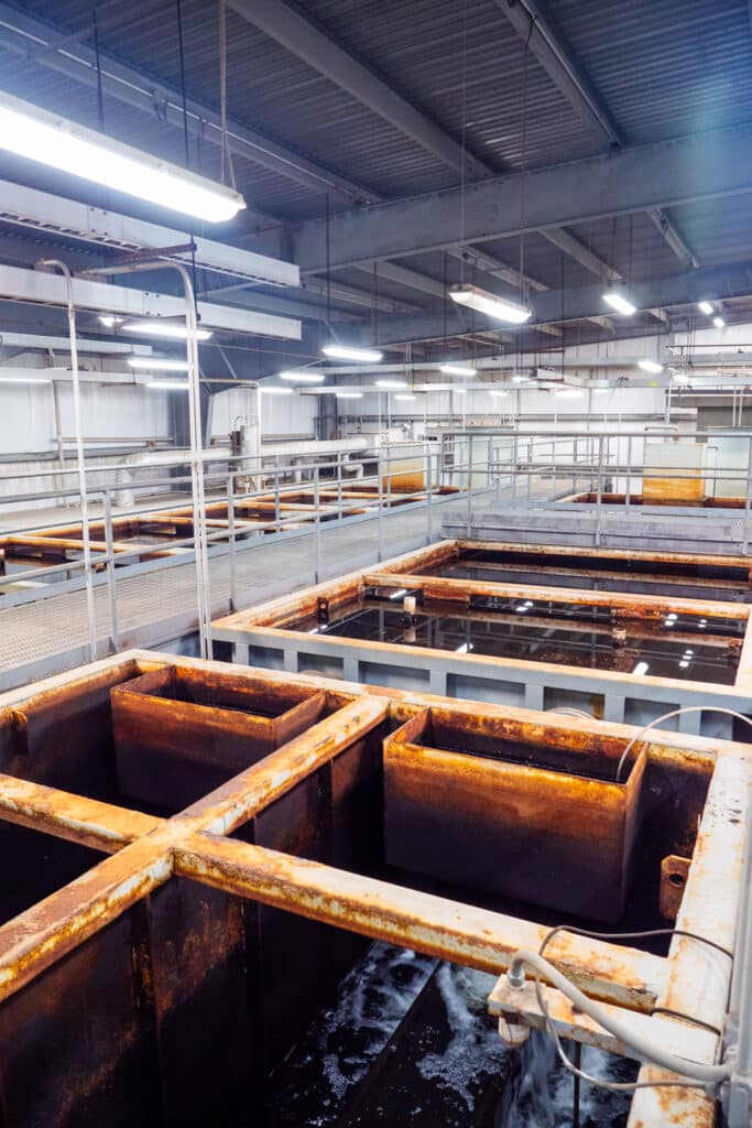A series of rusty rectangular metal tanks covering the floor of a Water Tech water treatment plant. A gray walkway threads through the tanks.