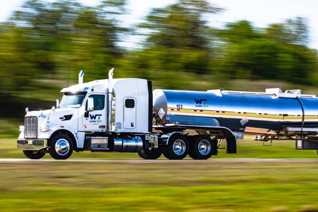 A shiny Water Tech transport truck driving on the freeway. The trees and grass are a blur and the truck is in perfect focus.