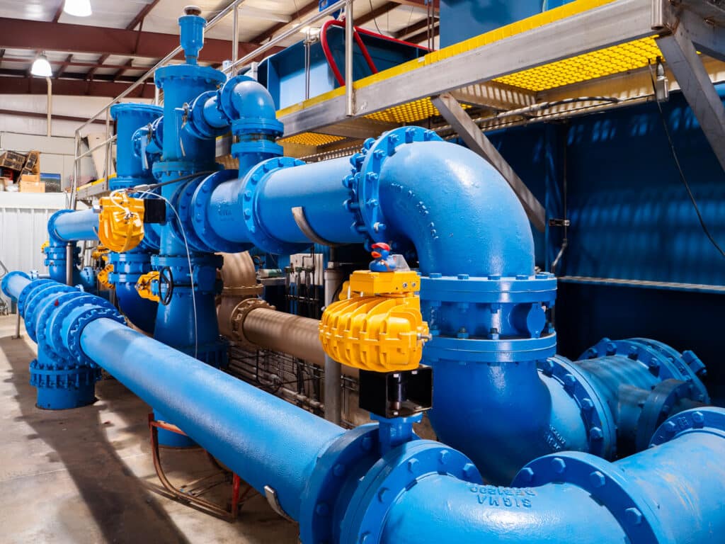 Large, bright blue pipes at the Water Tech wastewater treatment plant. The pipes are freshly painted blue and running above the pipes are bright yellow plastic shelves.